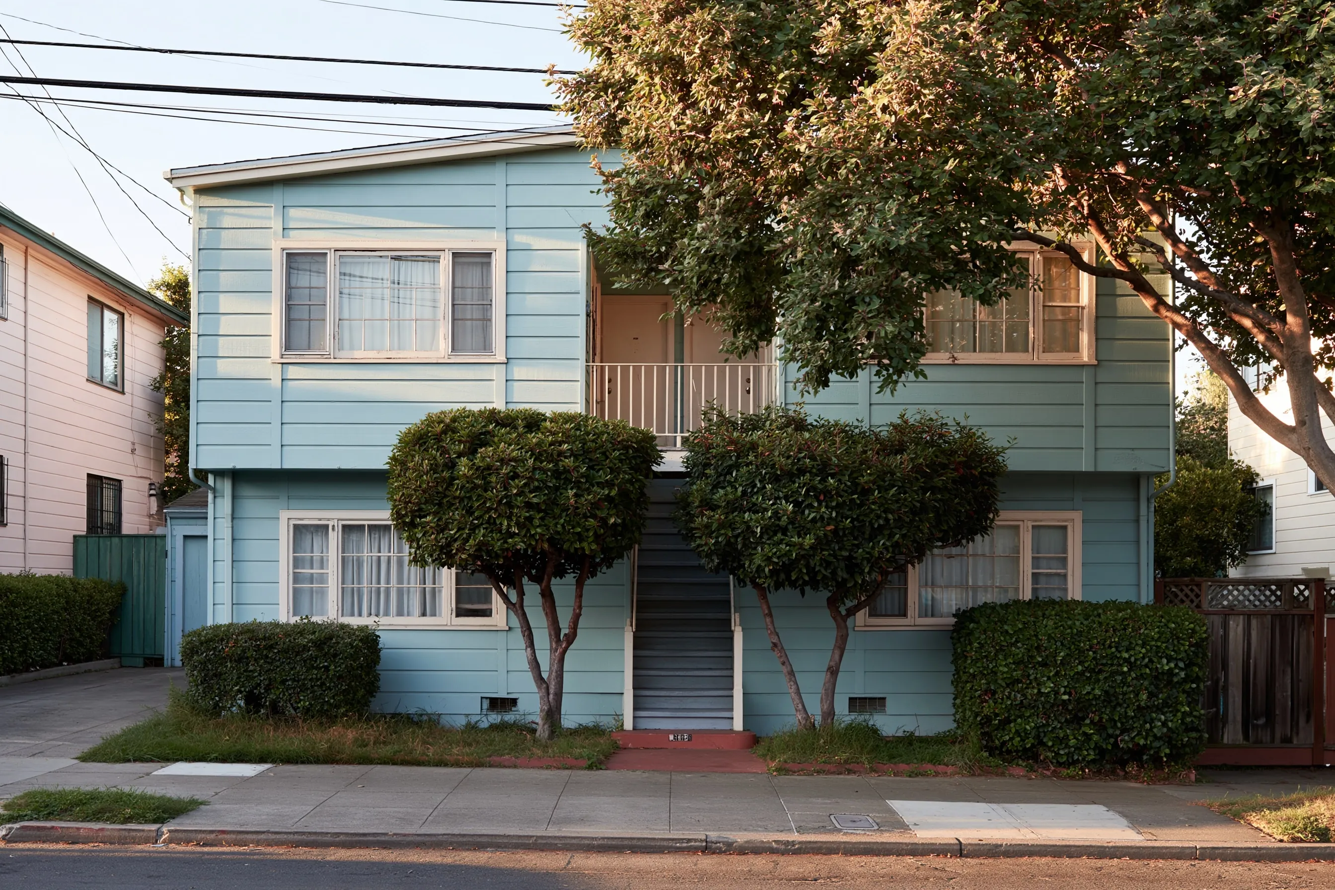 Two-story apartment building with painted wood siding, wood-framed windows and an exterior stair, two clipped trees in the front yard
